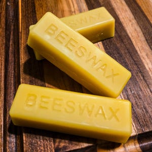 May include: Three rectangular bars of yellow beeswax, each embossed with the word BEESWAX in capital letters. The bars are arranged in a stack, showing a smooth, solid texture and a consistent golden-yellow color throughout each individual block.