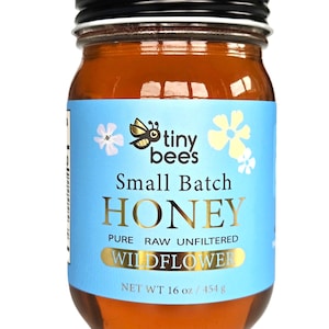 May include: A glass jar of wildflower honey with a black lid. The label is light blue with the text "tiny bees" and "Small Batch HONEY". The jar contains 16 oz / 454 g of pure, raw, unfiltered honey.