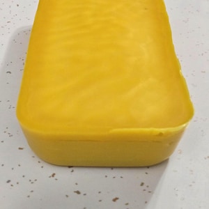 May include: A solid block of yellow beeswax, rectangular in shape with rounded edges. The wax has a smooth, slightly textured surface. The block is sitting on a white surface with small, dark flecks.