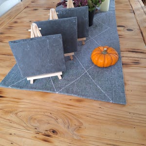 Könnte beinhalten: Drei kleine schwarze Schiefertafeln auf Holzstaffeleien, mit einem kleinen orangefarbenen Kürbis auf einem grauen Tischläufer mit weißem Rautenmuster. Der Tischläufer liegt auf einem Holztisch.