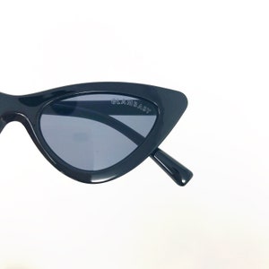 Puede incluir: Gafas de sol negras de ojo de gato con lentes grises oscuras. Las gafas tienen la palabra "GLAMBABY" impresa en el marco.