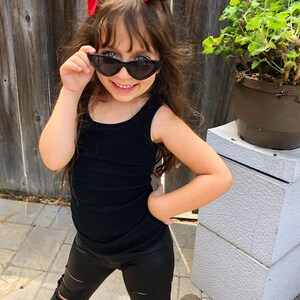 Puede incluir: Una ni&ntilde;a peque&ntilde;a con leggings negros rotos, un top negro sin mangas y gafas de sol negras de ojo de gato. Lleva un lazo rojo en el pelo.