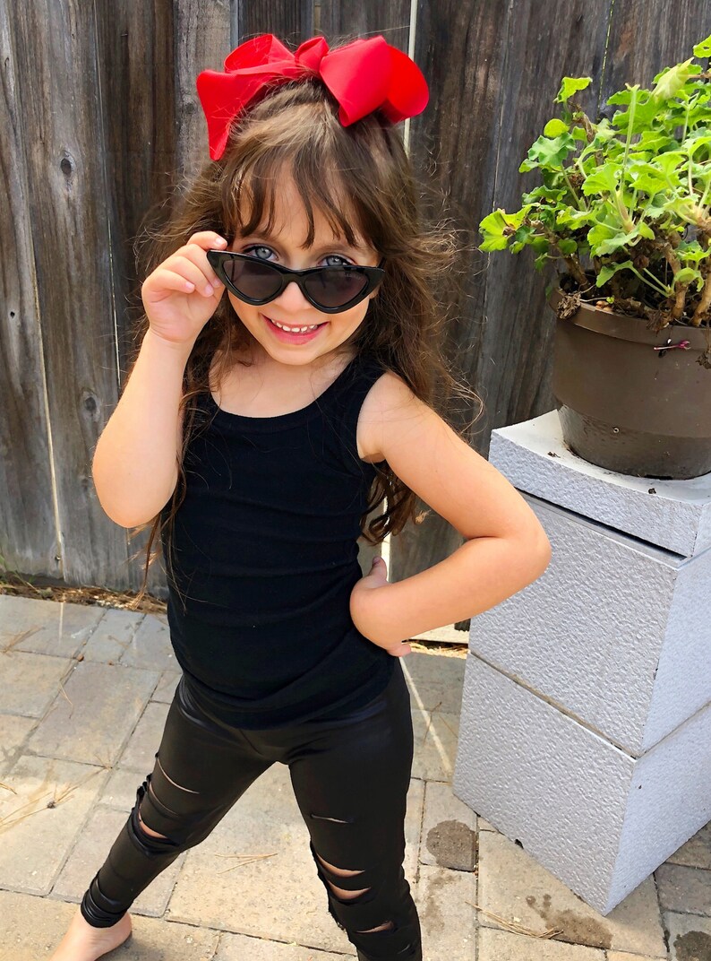 Puede incluir: Una ni&ntilde;a peque&ntilde;a con leggings negros rotos, un top negro sin mangas y gafas de sol negras de ojo de gato. Lleva un lazo rojo en el pelo.