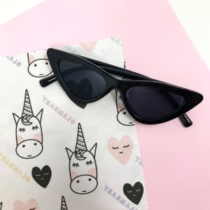 Puede incluir: Gafas de sol negras de ojo de gato con un paño de limpieza de microfibra blanco con un patrón repetido de unicornios y corazones. El paño tiene el texto "GLAMBABY" impreso.