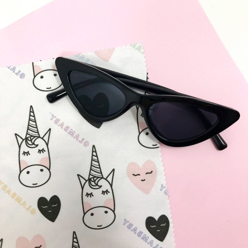 Puede incluir: Gafas de sol negras de ojo de gato con un pa&ntilde;o de limpieza de microfibra blanco con un patr&oacute;n repetido de unicornios y corazones. El pa&ntilde;o tiene el texto "GLAMBABY" impreso.