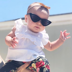 Puede incluir: Un beb&eacute; con un top blanco con volantes y leggings florales negros con gafas de sol negras de ojo de gato y una diadema marr&oacute;n con un lazo.
