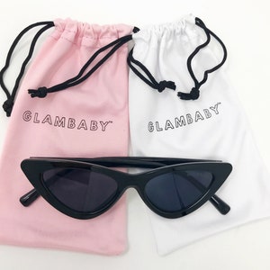 Puede incluir: Gafas de sol negras de ojo de gato con montura negra y lentes oscuros. Las gafas de sol est&aacute;n encima de dos bolsas de cord&oacute;n, una rosa y otra blanca, ambas con el texto "GLAMBABY" impreso.