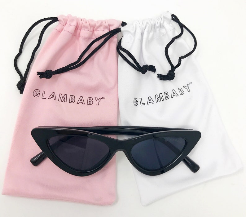 Puede incluir: Gafas de sol negras de ojo de gato con montura negra y lentes oscuros. Las gafas de sol est&aacute;n encima de dos bolsas de cord&oacute;n, una rosa y otra blanca, ambas con el texto "GLAMBABY" impreso.