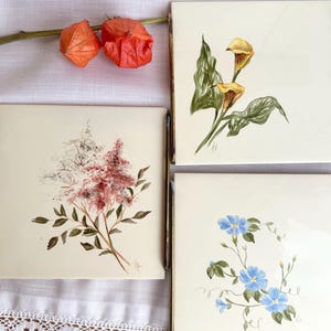 Może przedstawiać: Zestaw trzech kwadratowych płytek ceramicznych z motywami kwiatowymi. Jedna płytka przedstawia żółte kalie, druga czerwone i białe kwiaty, a trzecia niebieskie powoje. Każda płytka ma złotą metalową ramę.