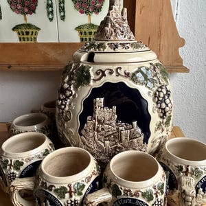 Può includere: Un set di boccali da birra in ceramica con un grande contenitore con coperchio e diverse tazze più piccole. Il set presenta un colore di base beige con accenti blu scuro e verdi, tra cui motivi di uva e castello. Le tazze hanno manici e testo decorativo.