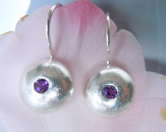 May include: A pair of silver drop earrings featuring a smooth, domed circular design with a brushed metallic finish. Each earring is centered with a small, round, faceted purple amethyst gemstone set securely into the silver dome.