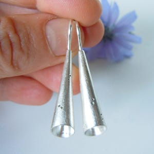 Könnte beinhalten: Ein Paar silberfarbene Ohrhänger in langer, schlanker, konischer Form mit gebürstetem, mattem Finish. Jeder Ohrring ist mit kleinen, verstreuten kreisförmigen Vertiefungen auf der Oberfläche versehen und verfügt über einen dünnen Haken.