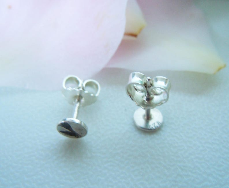 May include: A pair of silver stud earrings with butterfly backs. The flat, circular front faces are polished and reflective. The back of one earring is clearly stamped with the hallmark 925, indicating sterling silver composition.