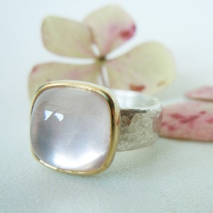 Könnte beinhalten: Ein silberner Ring mit gehämmerter Textur und einem quadratischen, klaren, hellrosa Edelstein, der in einer goldenen Fassung sitzt.