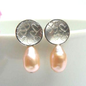 Op de afbeelding: Een paar oorbellen met een ronde, zilverkleurige metalen schijf voorzien van een herhalend hartjespatroon. Aan elke schijf hangt een enkele, gladde, peervormige, lichtroze parel, wat zorgt voor een gebalanceerd en symmetrisch ontwerp.