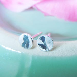 May include: A pair of circular stud earrings featuring a metallic finish with a dark, abstract botanical print on the front surface. Each earring has a standard post back mechanism for secure attachment, showcasing a flat, round design.