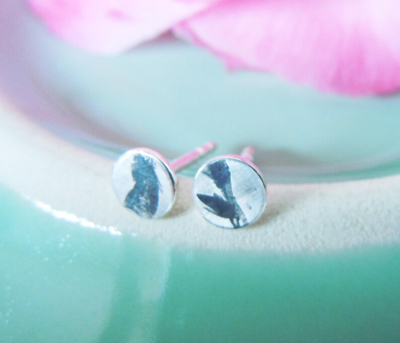 May include: A pair of circular stud earrings featuring a metallic finish with a dark, abstract botanical print on the front surface. Each earring has a standard post back mechanism for secure attachment, showcasing a flat, round design.