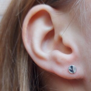 May include: A small, circular silver stud earring with a polished, reflective surface. The earring is worn in the earlobe, featuring a simple, flat, round design that catches the light, showcasing a smooth metallic finish and a clean, circular shape.