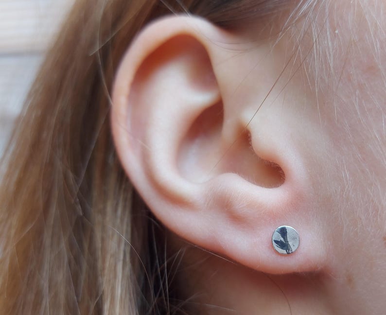 May include: A small, circular silver stud earring with a polished, reflective surface. The earring is worn in the earlobe, featuring a simple, flat, round design that catches the light, showcasing a smooth metallic finish and a clean, circular shape.