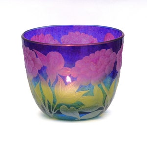 May include: A blue glass candle holder with a floral design. The flowers are pink and yellow and are etched into the glass. The candle holder is lit and the flame is visible.