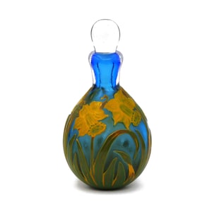 May include: A decorative glass perfume bottle with a clear stopper and a vibrant blue neck. The body features yellow daffodil floral designs on a teal background, with green leaves and stems. The bottle has a rounded shape.