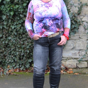 May include: A woman wearing a pink and white long-sleeved shirt with a black and white graphic print. The shirt has the text "The Best Of" printed on it. She is wearing blue jeans and black knee-high boots.