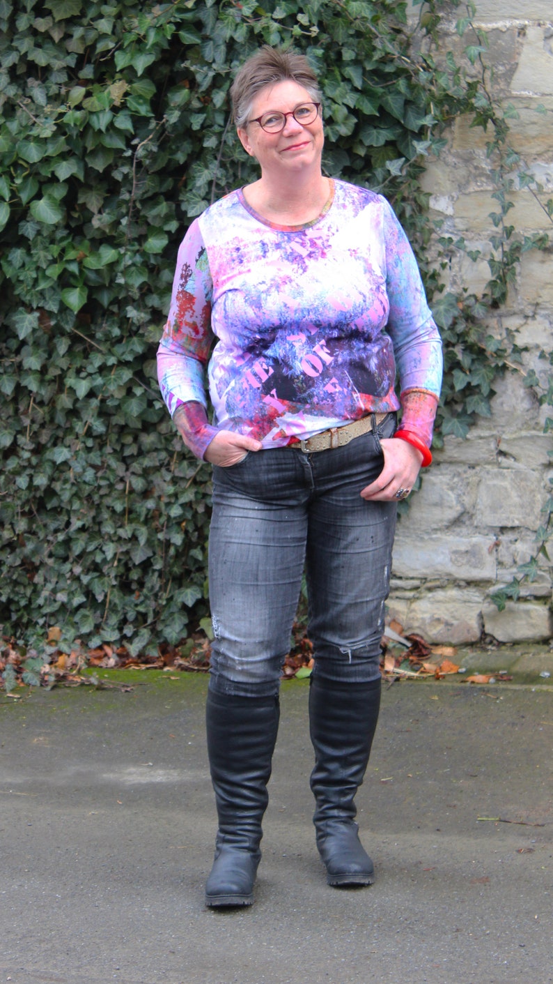 May include: A woman wearing a pink and white long-sleeved shirt with a black and white graphic print. The shirt has the text "The Best Of" printed on it. She is wearing blue jeans and black knee-high boots.
