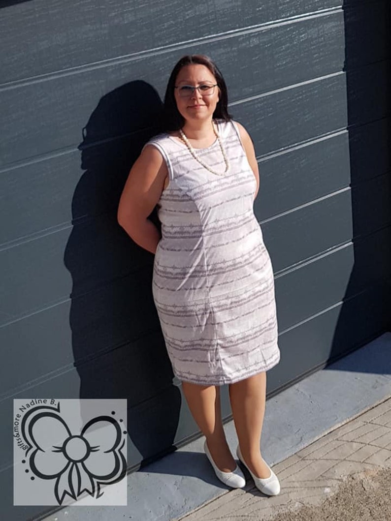 May include: A woman wearing a white and gray striped sleeveless dress with a pearl necklace and white shoes. She is standing in front of a gray garage door.