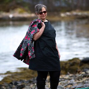 May include: A woman wearing a black dress with a floral print jacket draped over her shoulder. She is standing on a rocky shore with a body of water in the background.