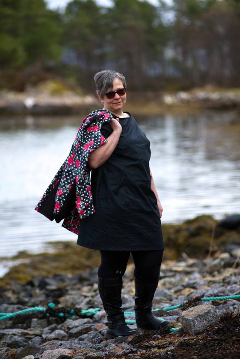 May include: A woman wearing a black dress with a floral print jacket draped over her shoulder. She is standing on a rocky shore with a body of water in the background.