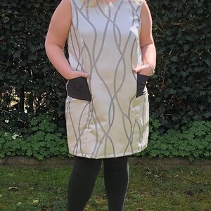 May include: A woman wearing a sleeveless dress with a gray and white abstract pattern. The dress has two black pockets on the front. She is wearing black leggings and gray ankle boots.