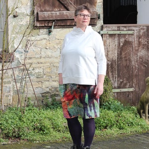 May include: A woman wearing a white long-sleeved shirt, a colourful patterned skirt, and black boots. She is standing in front of a stone building with a wooden door.
