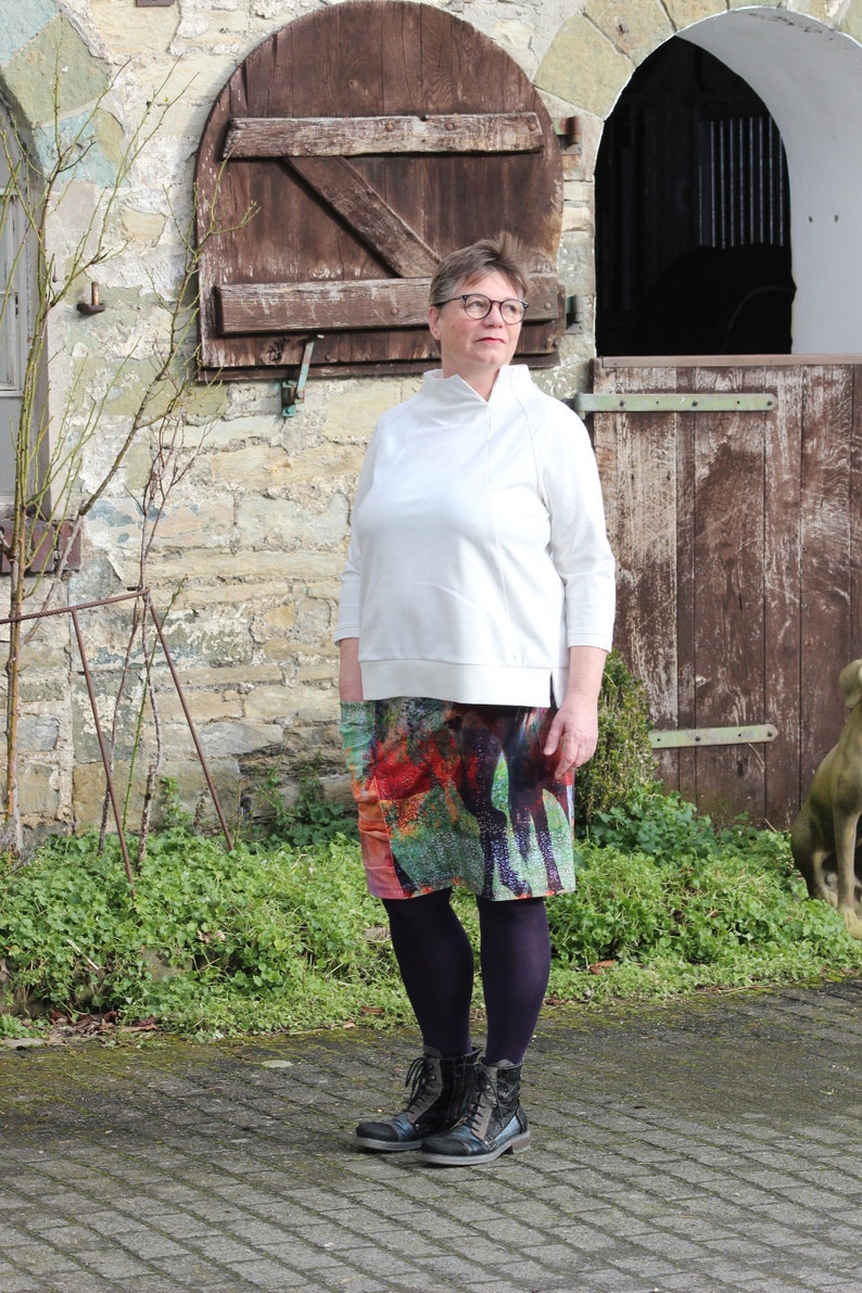 May include: A woman wearing a white long-sleeved shirt, a colourful patterned skirt, and black boots. She is standing in front of a stone building with a wooden door.