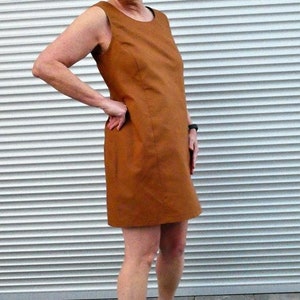 May include: A woman wearing a brown sleeveless dress and gray heels stands in front of a white slatted wall.