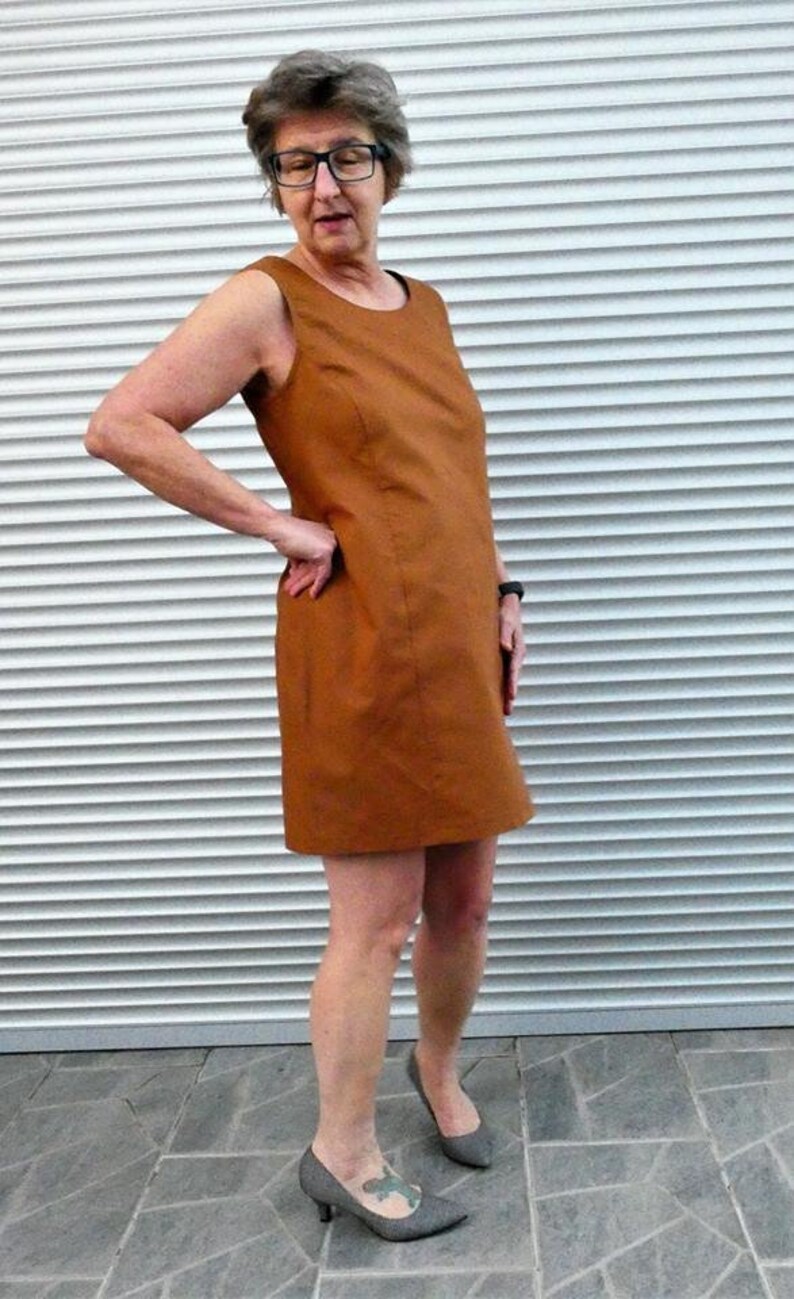 May include: A woman wearing a brown sleeveless dress and gray heels stands in front of a white slatted wall.