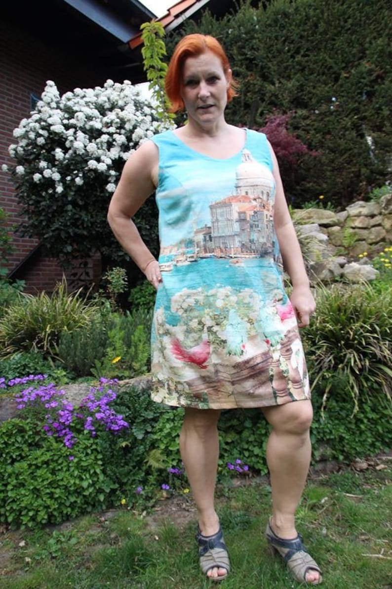 May include: A woman wearing a sleeveless dress with a printed image of a cityscape and a boat. The dress has a light blue background with a white and brown cityscape and a blue boat. The woman is standing in a garden with green grass and flowers.