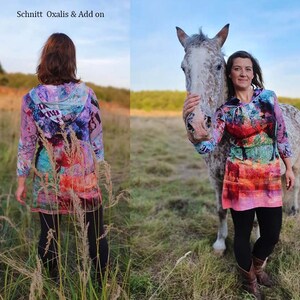 May include: A woman wearing a colourful hooded dress with abstract patterns stands in a field with a horse. The dress has a long sleeve and a loose fit. The woman is holding the horse's head with her left hand. The horse is white with brown spots. The background is a field of green grass.