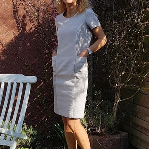 May include: A woman wearing a light gray dress with short sleeves and pockets. She is standing in a garden with a white wooden chair and green grass.