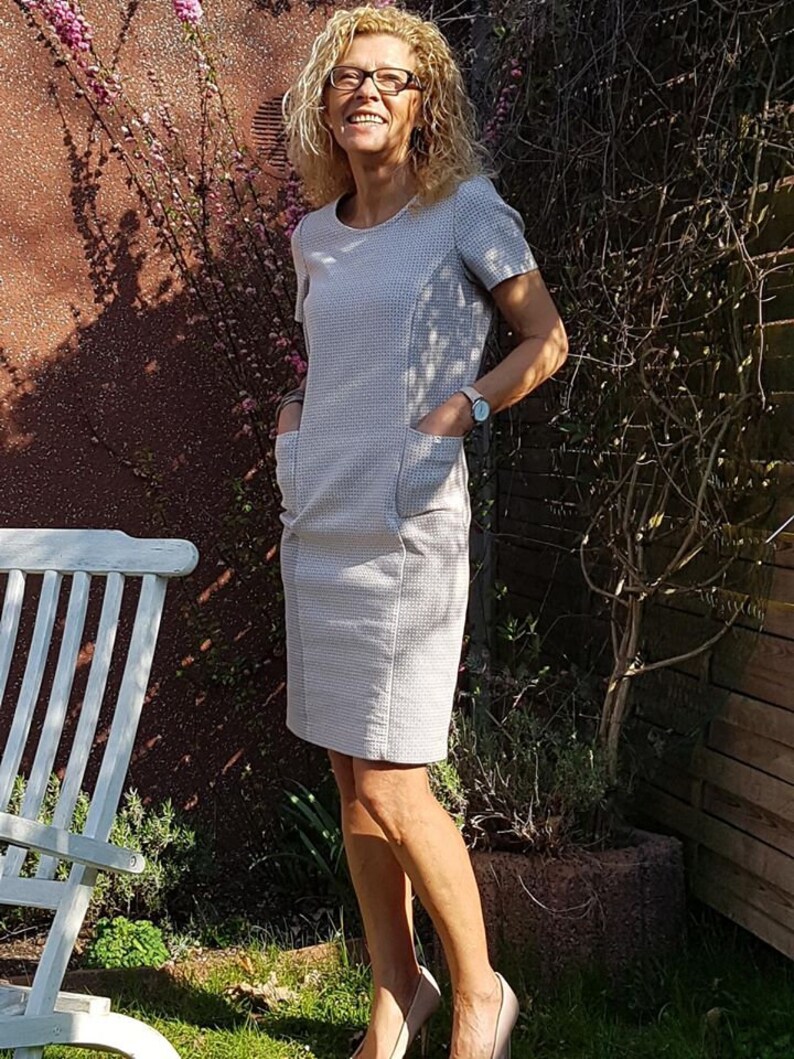 May include: A woman wearing a light gray dress with short sleeves and pockets. She is standing in a garden with a white wooden chair and green grass.