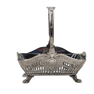 Könnte beinhalten: Ein silberfarbener Metallkorb mit einem verzierten, durchbrochenen Gitterdesign mit Blumen- und Blattmotiven. Der Korb hat einen hohen, gebogenen Griff, einen rechteckigen Körper und steht auf vier detaillierten klauenartigen Füßen.