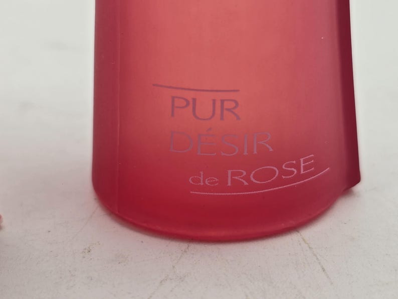 May include: A close-up of a red bottle with the words "PUR D&Eacute;SIR de ROSE" printed in a lighter color. The bottle has a smooth, matte finish and a slightly curved shape. The text is centered on the bottle.