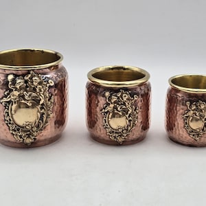 Może przedstawiać: Zestaw trzech metalowych donic w kolorze miedzi o młotkowanej fakturze. Każda donica ma złoty brzeg i ozdobny złoty herb z cherubinami. Donice są różnej wielkości, tworząc schodkową ekspozycję.