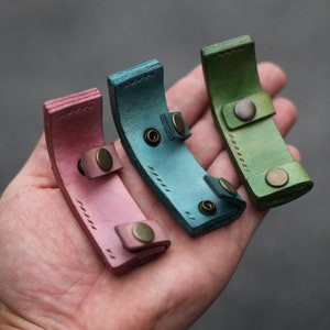 Könnte beinhalten: Drei Leder-Schlüsselanhänger in verschiedenen Farben: rosa, blau und grün. Jeder Schlüsselanhänger hat eine kleine Metallschnalle und Nähdetails.