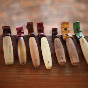Puede incluir: Siete cuchillos de talla con mango de madera y vainas de cuero en varios colores, incluyendo rojo, verde, amarillo y marrón. Los cuchillos están dispuestos en una fila sobre una superficie de madera.