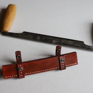 Puede incluir: Una funda de cuero marrón con costuras y broches metálicos para una herramienta de talla de madera.