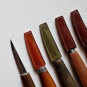 Puede incluir: Cinco cuchillos de talla envueltos en cuero con mangos de madera. Los cuchillos tienen diferentes envolturas de cuero de colores: marrón, naranja, verde y dos tonos de rojo.