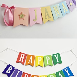 May include: A pastel-colored banner spelling JANA with gold glitter letters and stars, tied with pink ribbon, and a separate rainbow-colored banner with white letters spelling HAPPY BIRTHDAY, both featuring swallowtail pennant flags on white string.