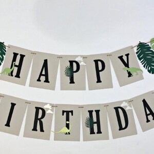 K&ouml;nnte beinhalten: Ein Banner mit den Worten "Happy Birthday" in schwarzen Buchstaben auf beigefarbenem Hintergrund. Das Banner ist mit gr&uuml;nen Bl&auml;ttern und Dinosauriern verziert.