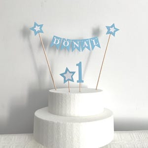 May include: A light blue cake topper set featuring a bunting banner with the name DONAL in white letters, two star-shaped toppers, and a number 1 topper. The decorations are attached to thin wooden sticks and are displayed on a white cake dummy.