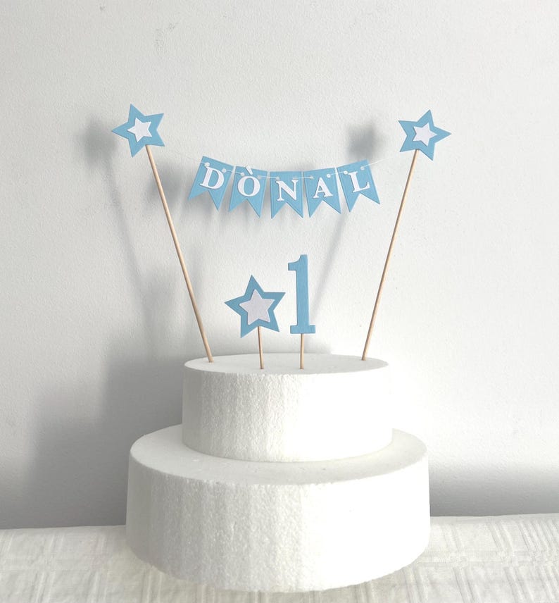 May include: A light blue cake topper set featuring a bunting banner with the name DONAL in white letters, two star-shaped toppers, and a number 1 topper. The decorations are attached to thin wooden sticks and are displayed on a white cake dummy.